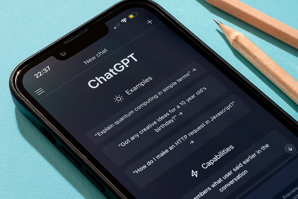 Pantalla de un teléfono móvil mostrando la interfaz de ChatGPT con ejemplos de preguntas, sobre un fondo azul claro junto a dos lápices de madera.