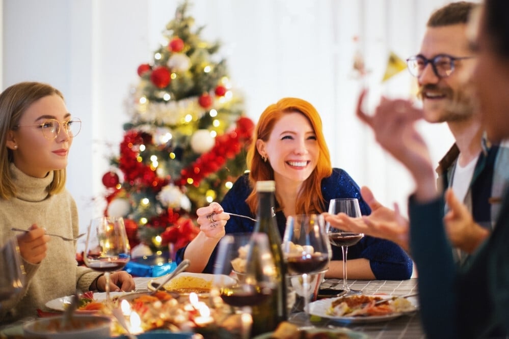 Mesa navideña con platos variados y copas de vino, personas compartiendo comida en un ambiente festivo con árbol decorado y luces en el fondo.