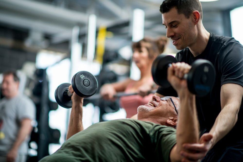 Un entrenador personal ayuda a una persona de la tercera edad durante un entrenamiento con mancuernas en un gimnasio.