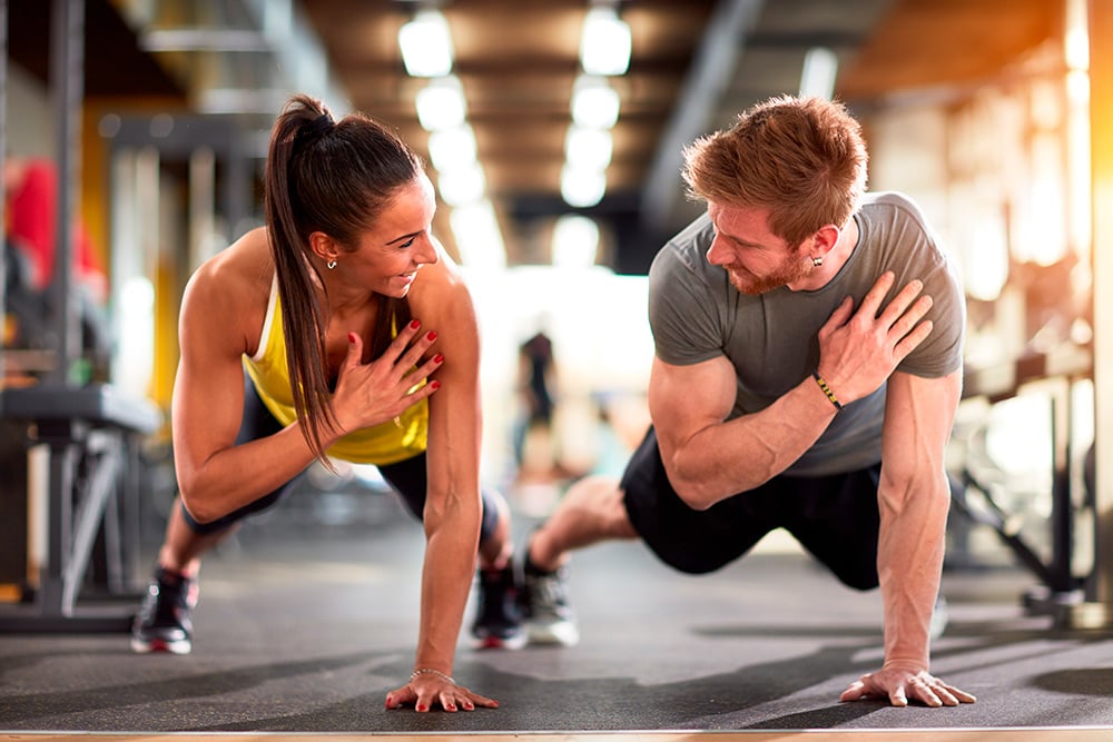 Hombre y mujer sonrientes realizan una plancha lateral en un gimnasio, apoyando una mano en el pecho mientras mantienen la postura y se motivan mutuamente.