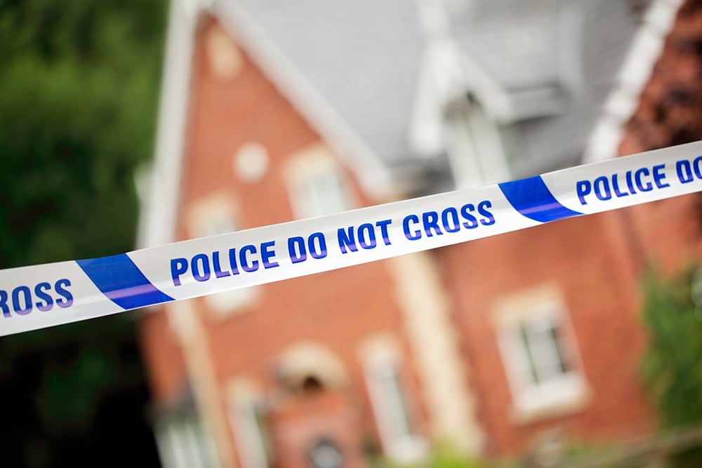 Cinta policial con el mensaje “Police Do Not Cross” delimitando el acceso frente a una vivienda de ladrillo desenfocada en el fondo.
