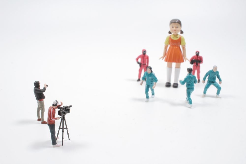 Miniaturas recreando una escena icónica con una muñeca gigante y personajes vestidos con uniformes verdes y rojos, mientras dos figuras simulan grabar con cámara.