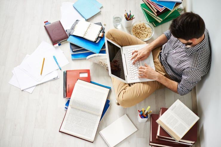 Un estudiante sentado en el suelo y rodeado de apuntes y libros abiertos escribe en un portátil.