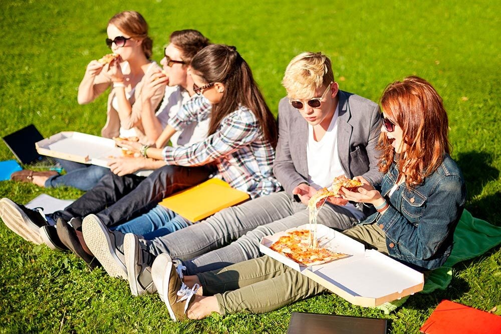 Dos chicas y tres chicos sentados en el césped compartiendo pizzas en un día soleado, disfrutando de un descanso universitario