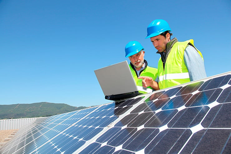 Dos ingenieros con casco azul consultan un portátil colocado sobre una placa solar. 