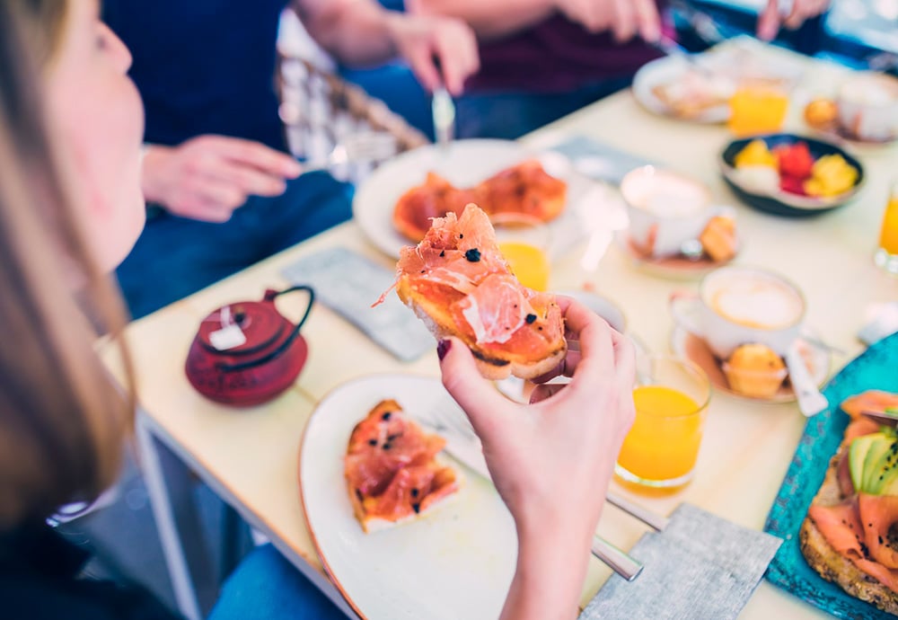 Imagen en la que se ve  una mesa con distintos platos de desayuno y l mano de una mujer que sostiene una tosta con jamón. 