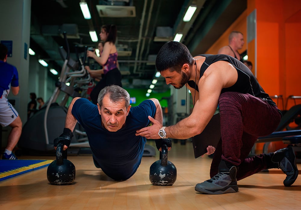 Un monitor en un gimnasio instruye a un hombre mayor que realiza un ejercicio en el suelo