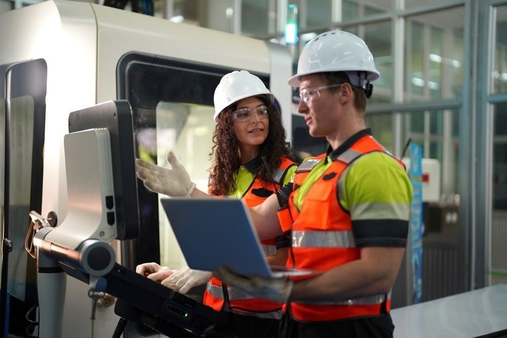 Dos personas con chalecos reflectantes y cascos blancos trabajando frente a una máquina industrial, usando un portátil y una pantalla táctil.