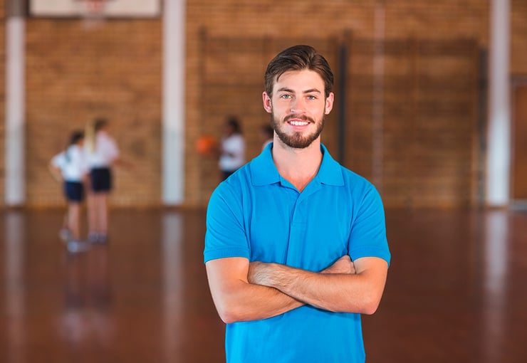 Profesor de Educación Física en un gimnasio escolar con brazos cruzados mirando a la cámara. 