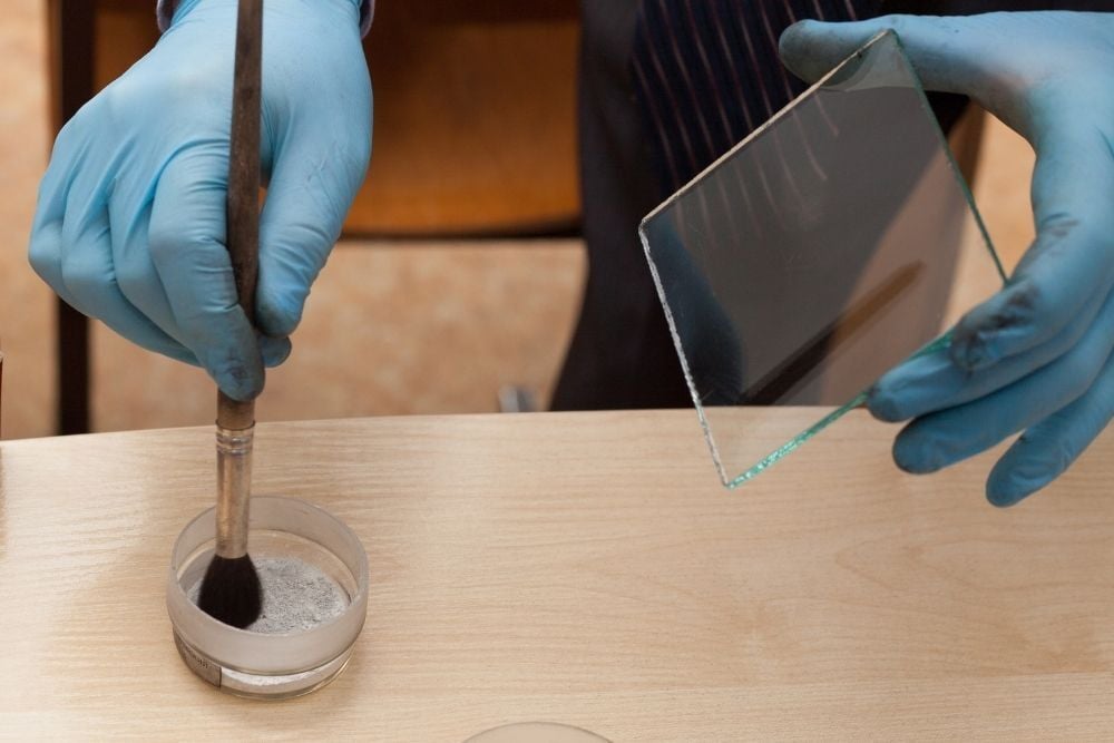 Técnico forense utilizando polvo para revelar huellas dactilares sobre una superficie de cristal