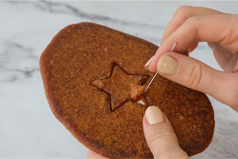 Manos sosteniendo una galleta marrón con forma de estrella, utilizando una aguja para recortar la figura.