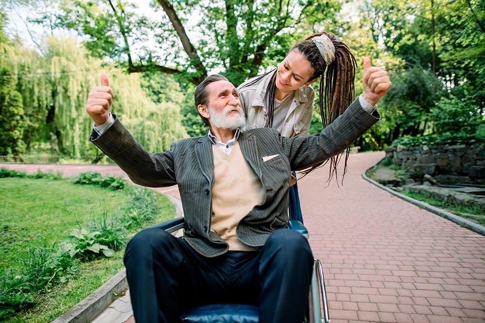 Una mujer joven con rastas empuja sonriendo la silla de ruedas de un hombre mayor feliz con los pulgares levantados. 