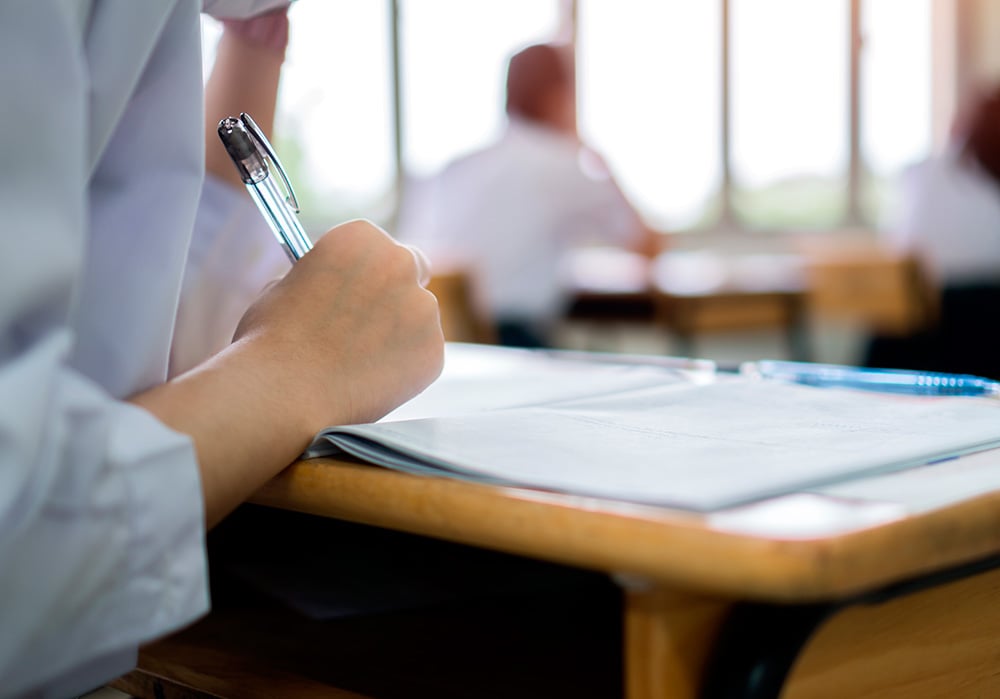 Una fotografía muestra la mano de una estudiante sujetando un bolígrafo mientras realiza un examen.