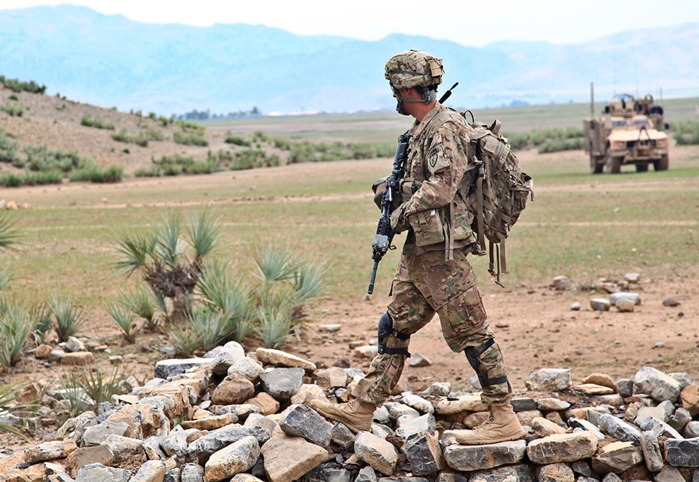 Soldado estadounidense durante una patrulla por Afganistán y al fondo un blindado y unas montañas.  
