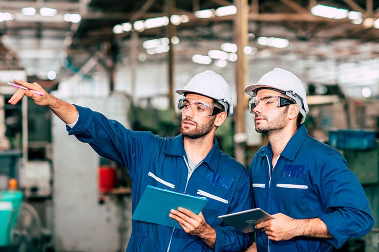 Dos técnicos de prevención con mono azul y equipados con casco y gafas de seguridad, supervisan una fábrica.