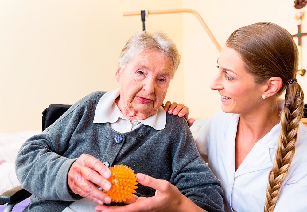 Anciana en silla de ruedas sostiene una pelota de pinchos y a su lado una terapeuta ocupacional la mira sonriendo.