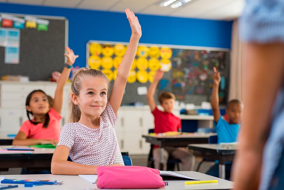 Niña, junto a otros compañeros de clase, sentada y levantando la mano.