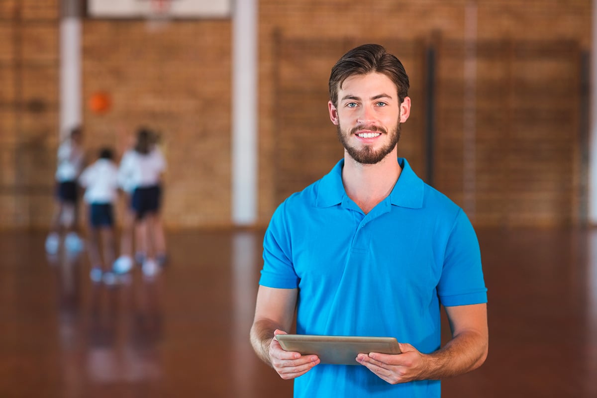 Profesor de educación física supervisando a niños en un gimnasio, utilizando una tablet para guiar la actividad