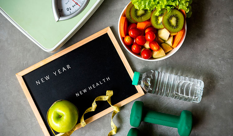 Imagen alegórica de los propósitos de año nuevo en el que se ve una manzana, una báscula, una cinta métrica, un cuenco con fruta y una pizarra en al que se puede leer en inglés: 
