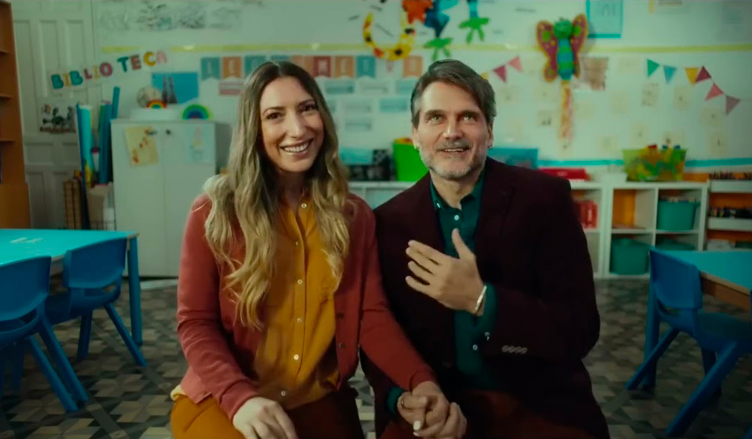 Una mujer y un hombre sentados juntos en una clase infantil, sonrientes y cogidos de la mano. Detrás de ellos se ven mesas y sillas de colores, dibujos infantiles en la pared y decoración escolar. La escena forma parte del nuevo anuncio navideño de Campofrío.