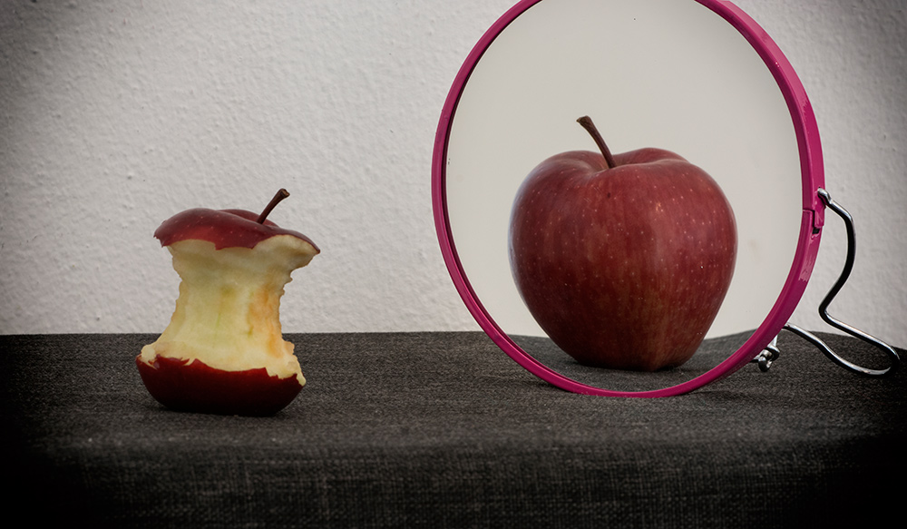 Una manzana mordida delante de un espejo en el que el reflejo muestra una manzana entera como un simbolismo sobre lo que significan los trastornos de la alimentación.