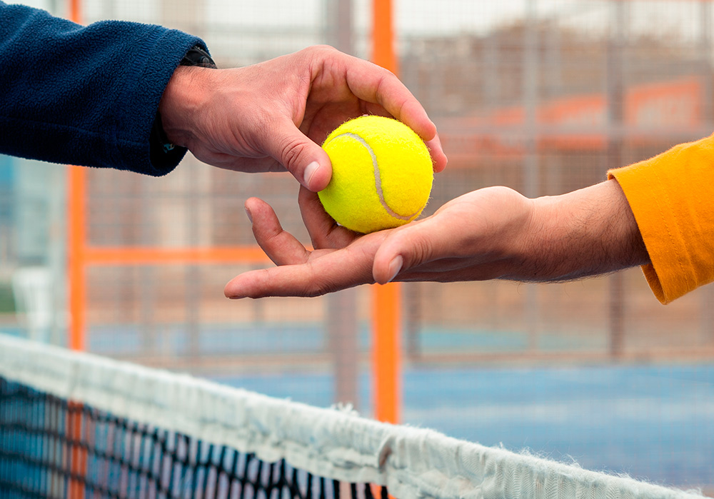 La mano de un jugador de pádel entrega la pelota a otro jugador por encima de la red de la cancha.
