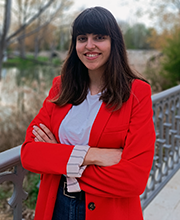 "Paula Herrero, con una americana roja y los brazos cruzados, posa sobre un puente con un río de fondo.