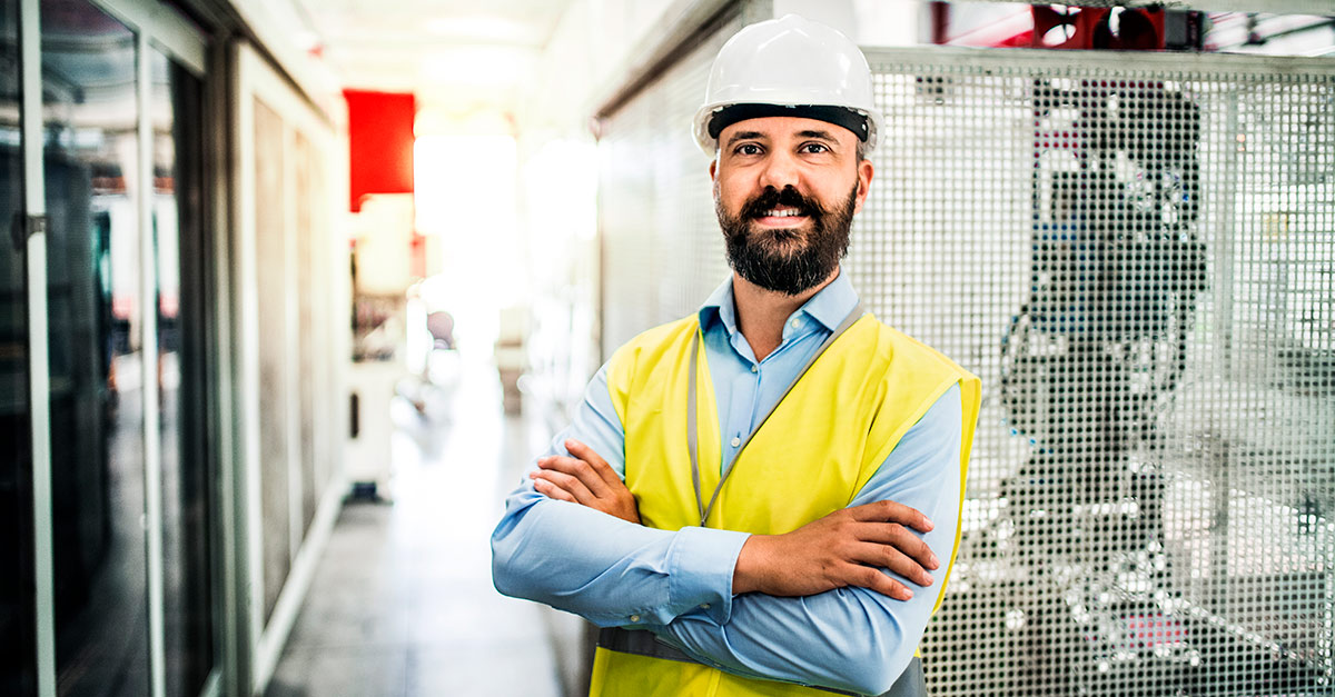 Un joven ingeniero de organización industrial con barba y equipado con casco blanco y chaleco reflectante con los brazos cruzados mirando a cámara. 